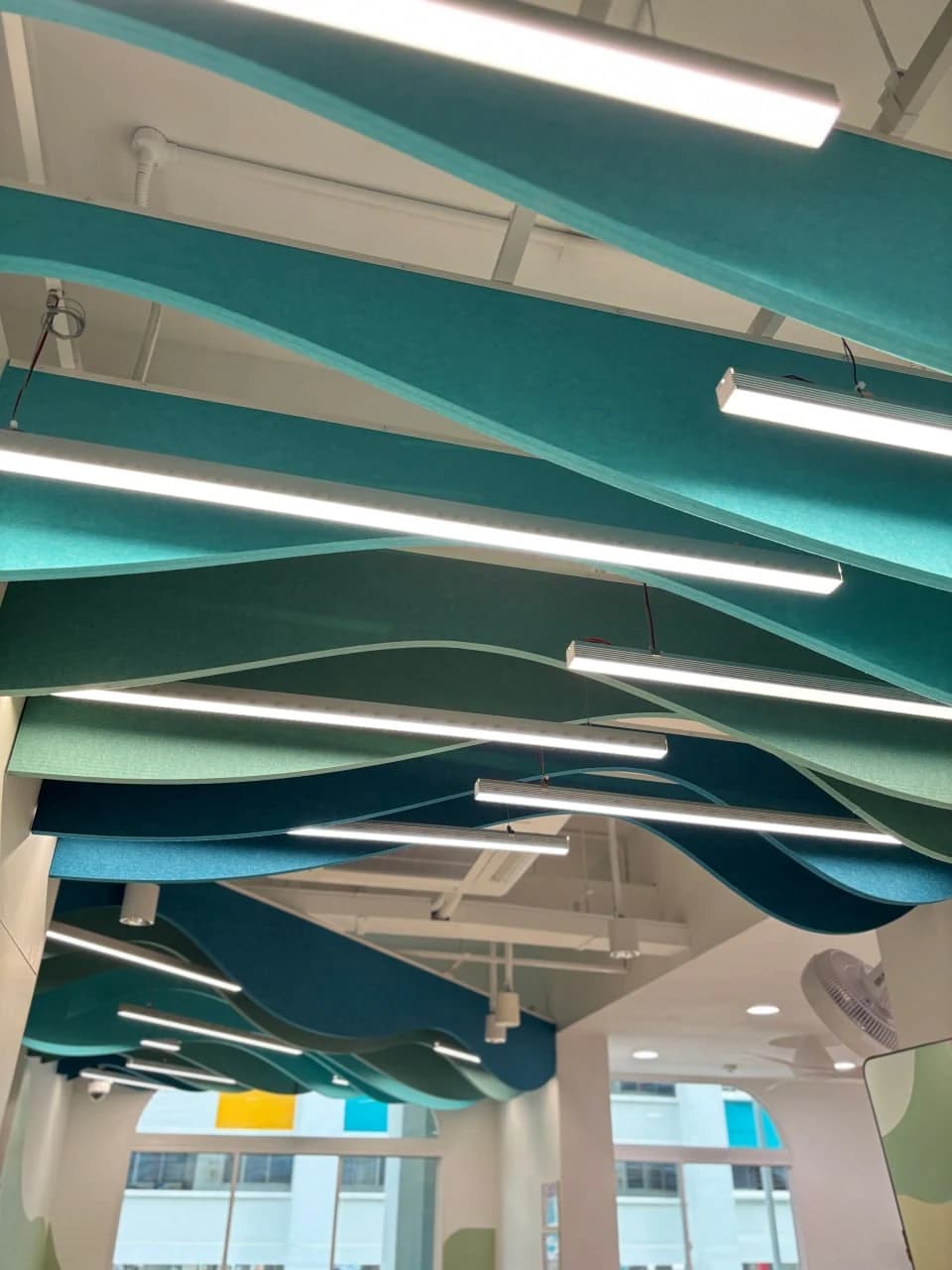 Layered teal wave acoustic ceiling panels with integrated linear lighting at NTUC Health Marsiling